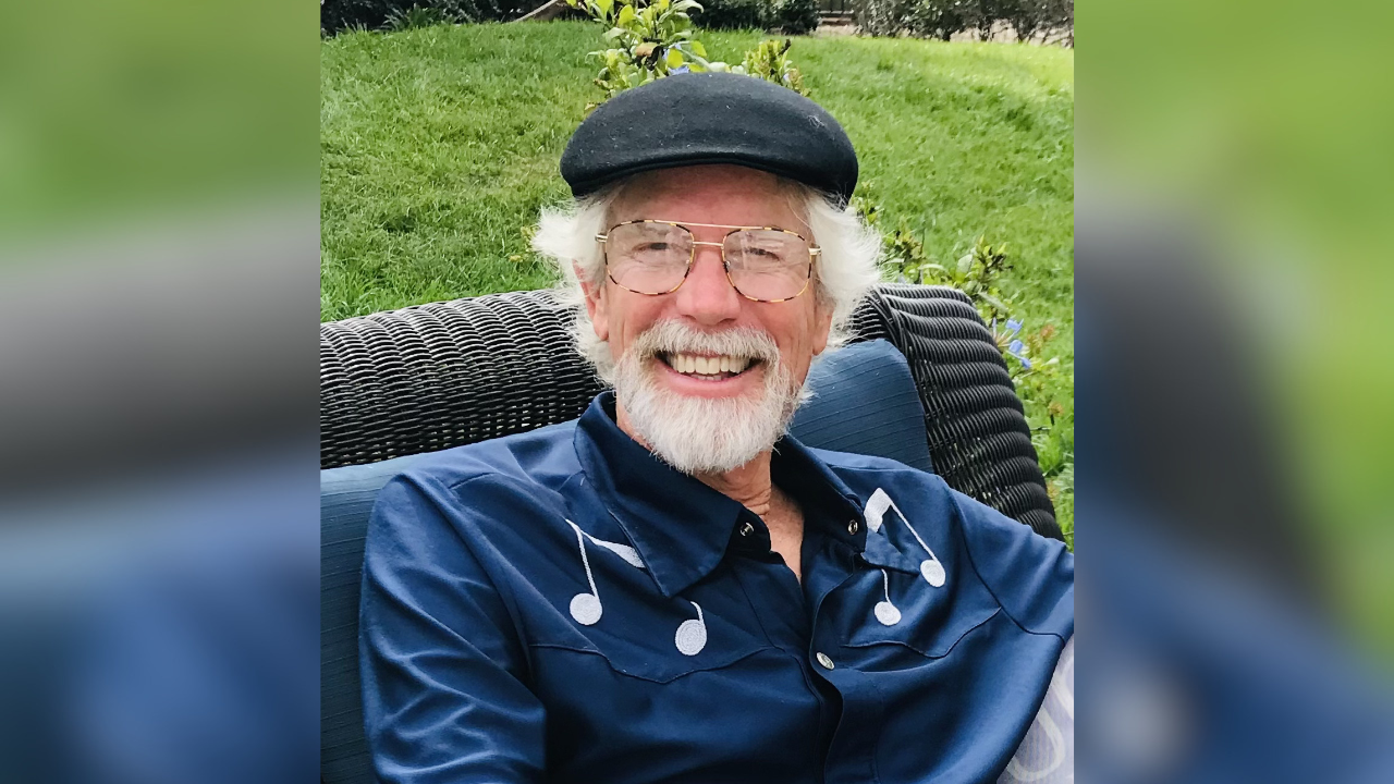 Echoes of Jazz host Rusty Barker, a smiling man in a flat cap and blue shirt with musical notes